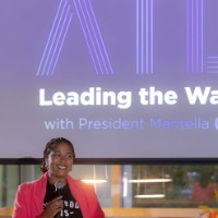 Woman wearing pink blazer smiling as she hold microphone and phone